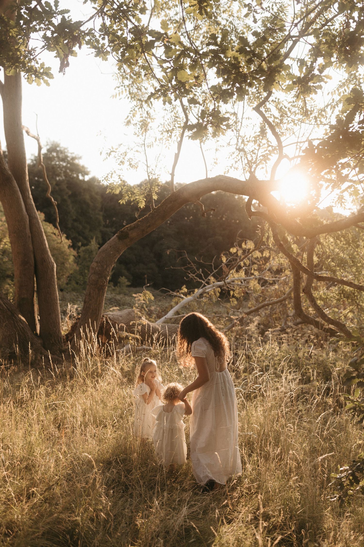 Two figures in white dresses dance through a sunlit meadow with tall grass and trees at golden hour.