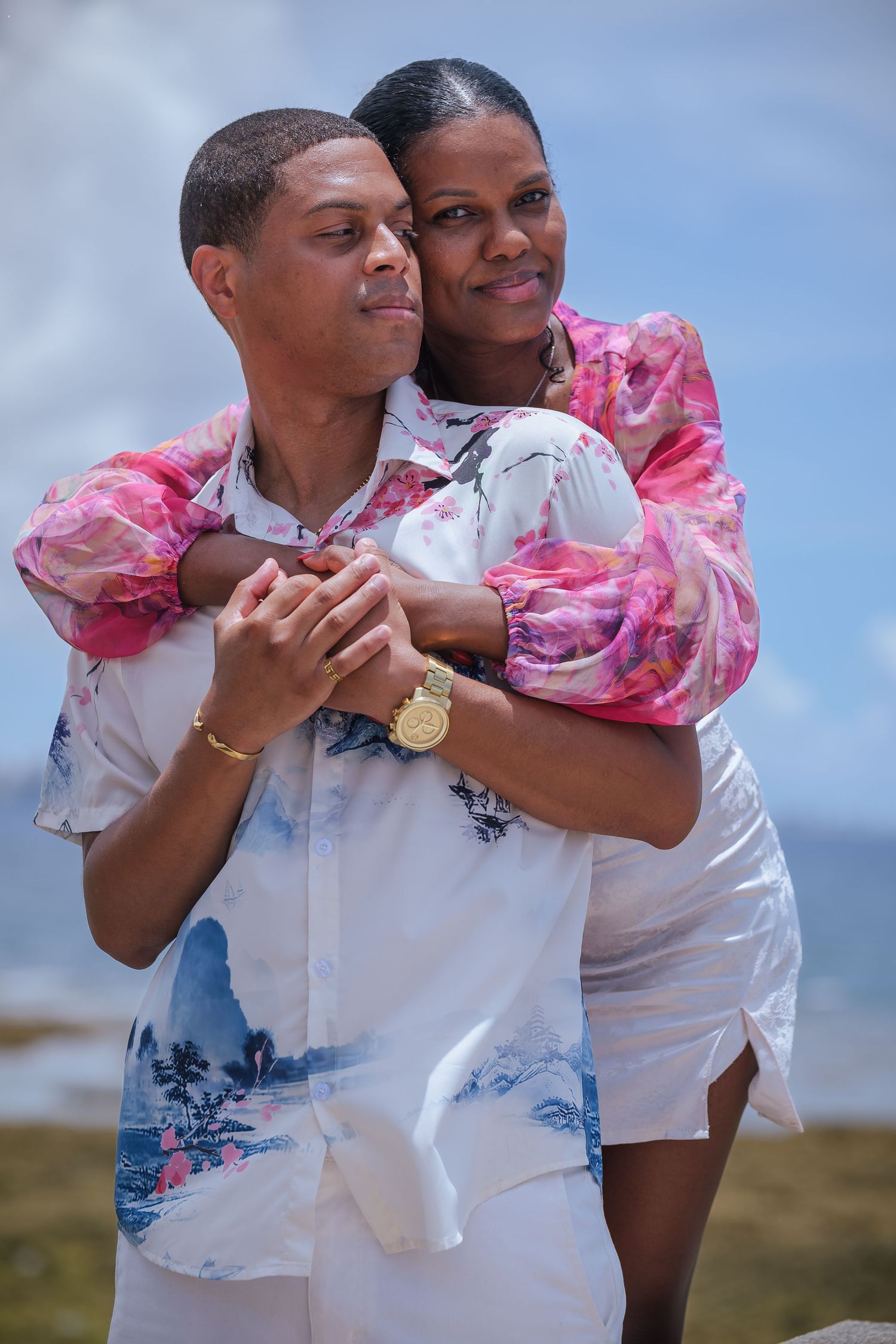 A couple embraces outdoors wearing coordinated pink and white outfits against a blue sky background.