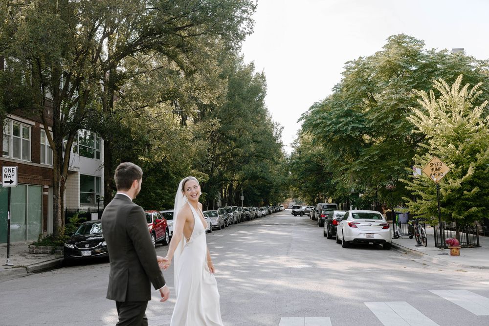A couple in wedding attire walks down a tree-lined street in an urban neighborhood during a photo session.