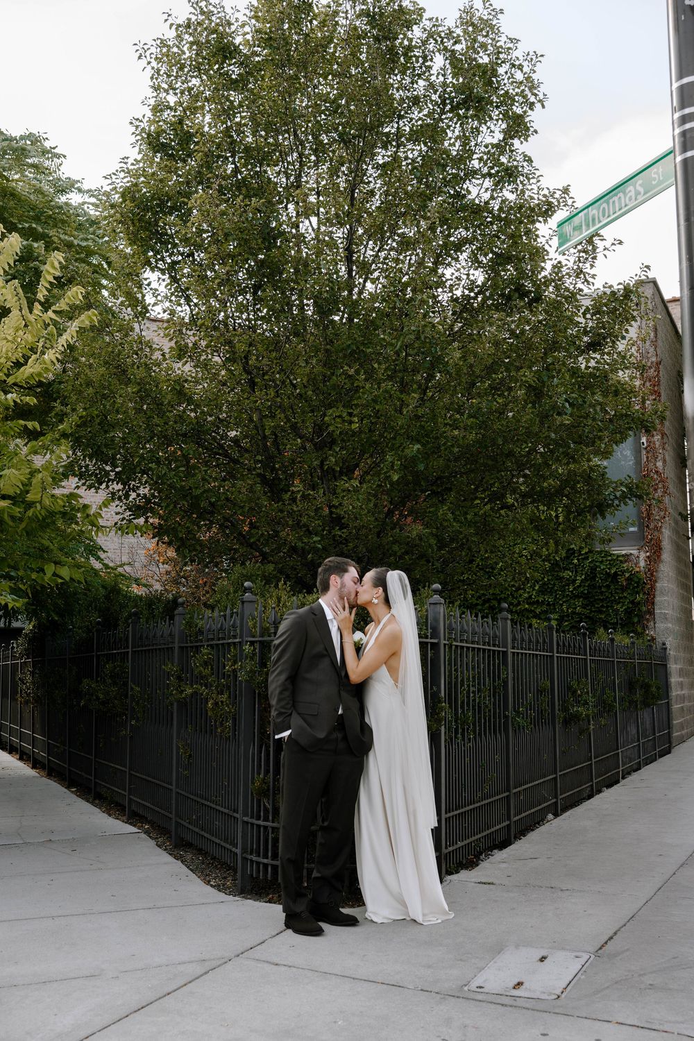 A wedding couple poses against a black iron fence and trees in an urban setting.