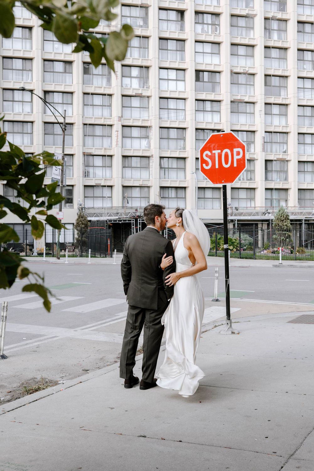 A couple in wedding attire shares a romantic moment near a stop sign outside a modern office building.