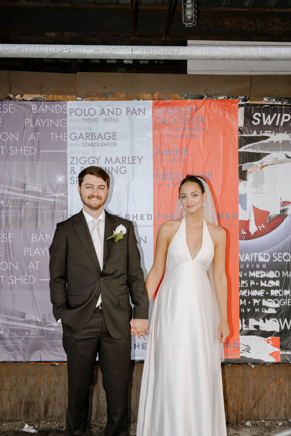 A couple in wedding attire poses against a wall covered in colorful posters and graffiti.