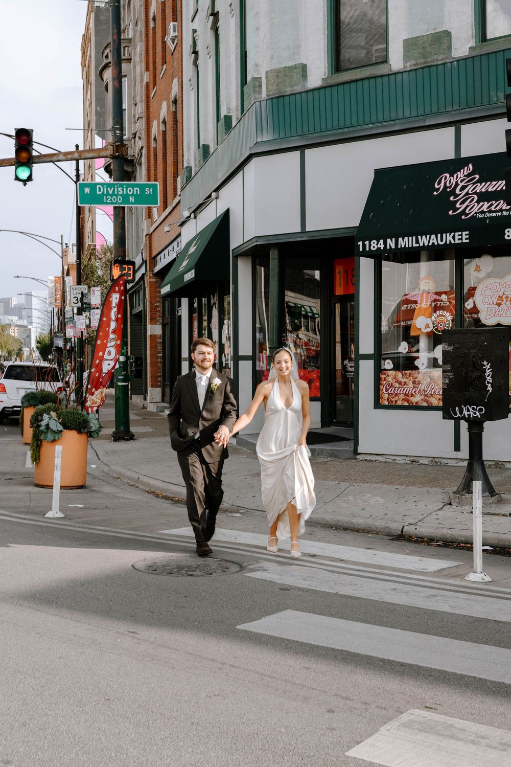 A couple in wedding attire walks together on a city street with brick buildings and storefronts in the background.
