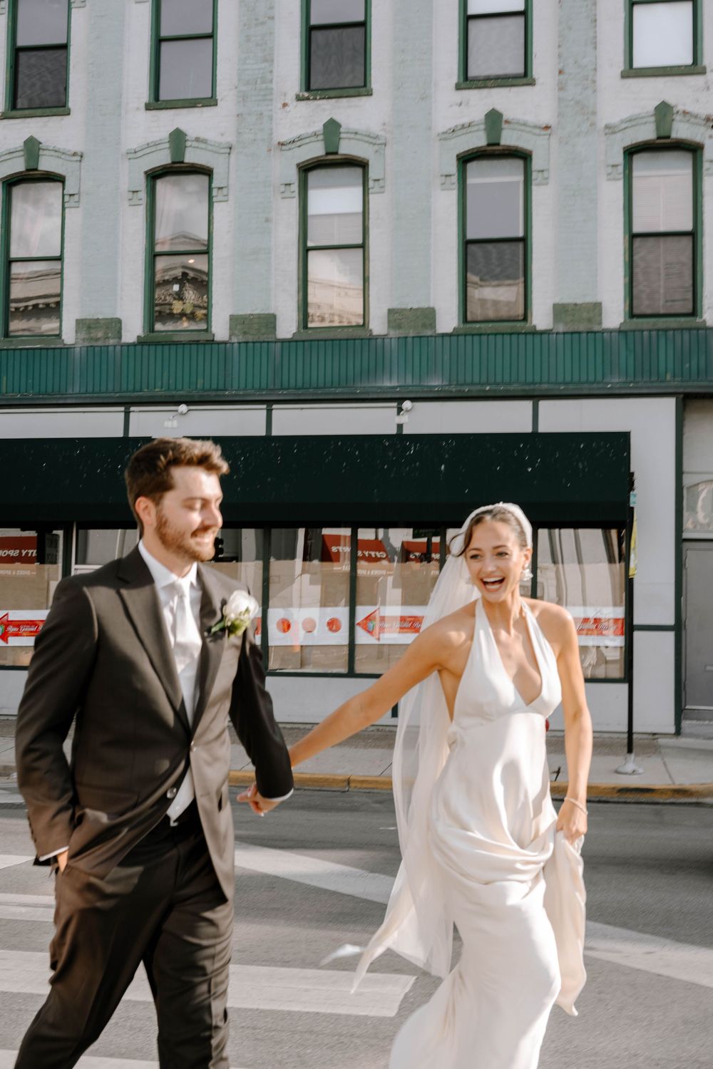 A couple in wedding attire runs playfully in front of a white building facade.