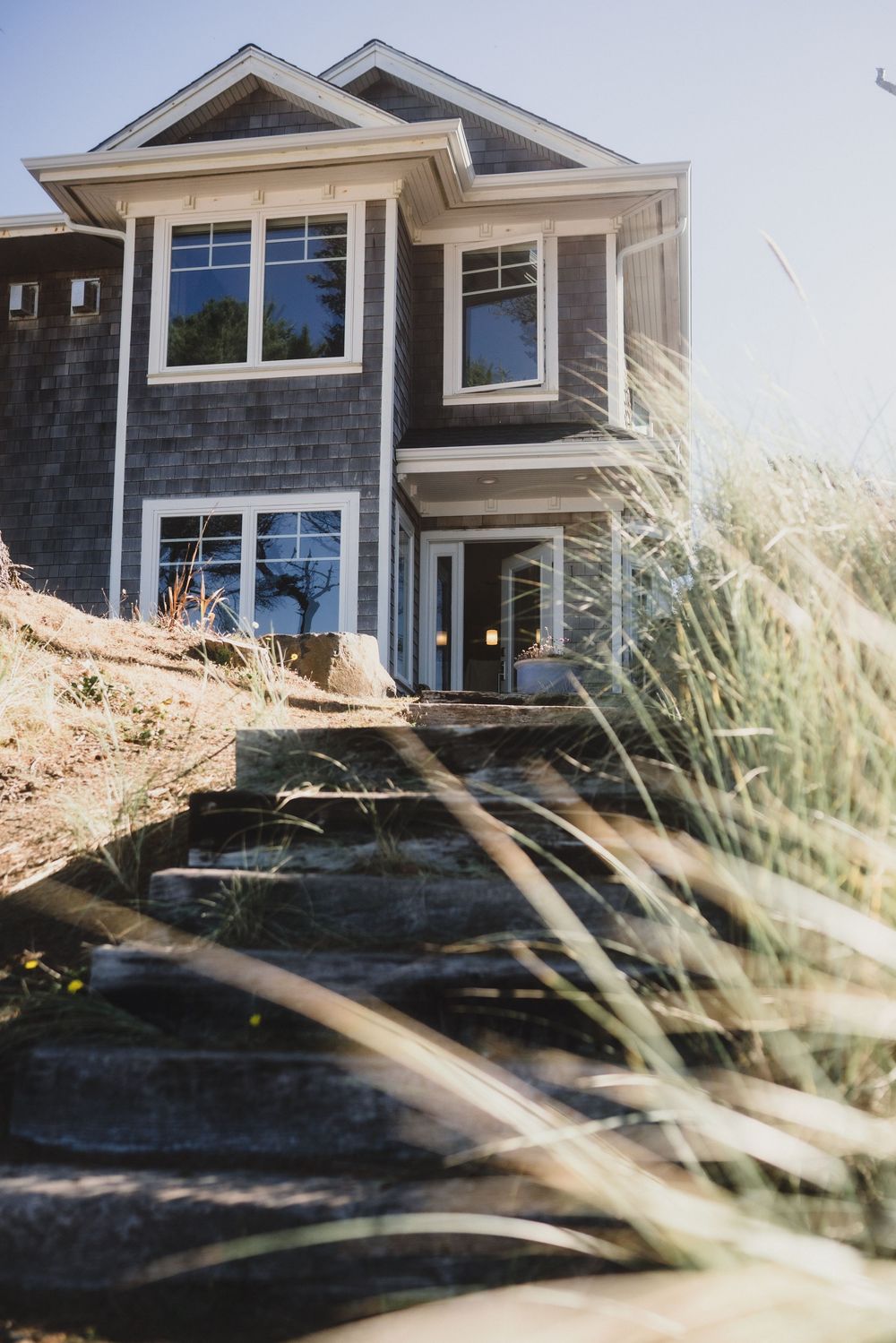 Modern craftsman style house with grey siding and white trim viewed through swaying beach grass in sunlight.