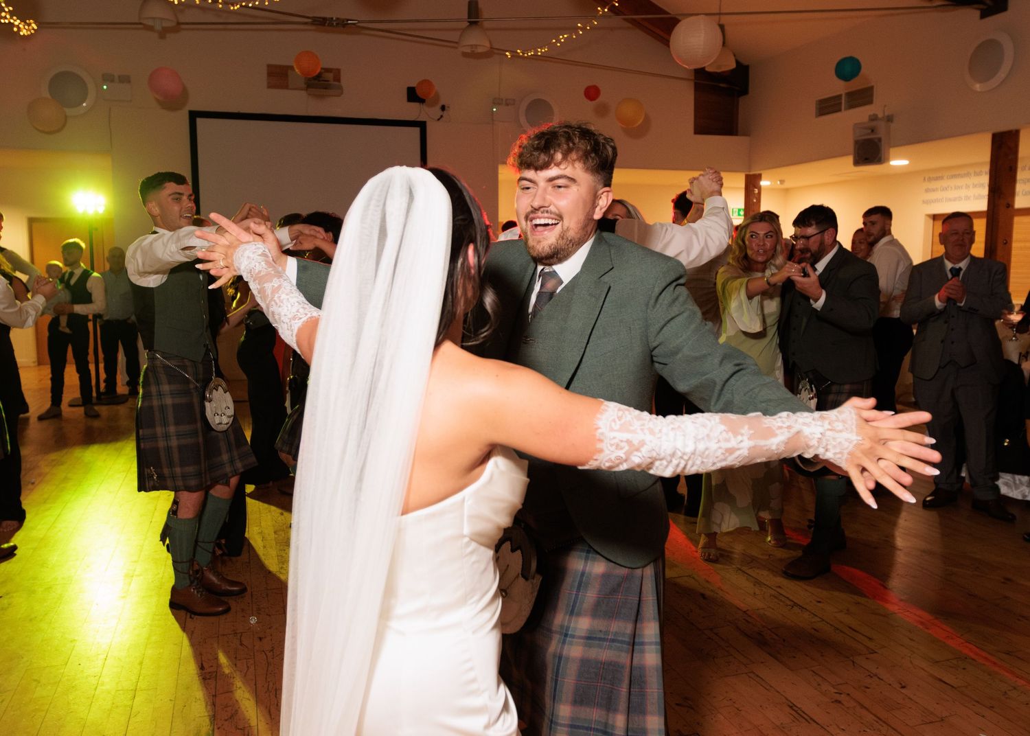 Wedding celebration with string lights overhead as guests dance in a large indoor venue.