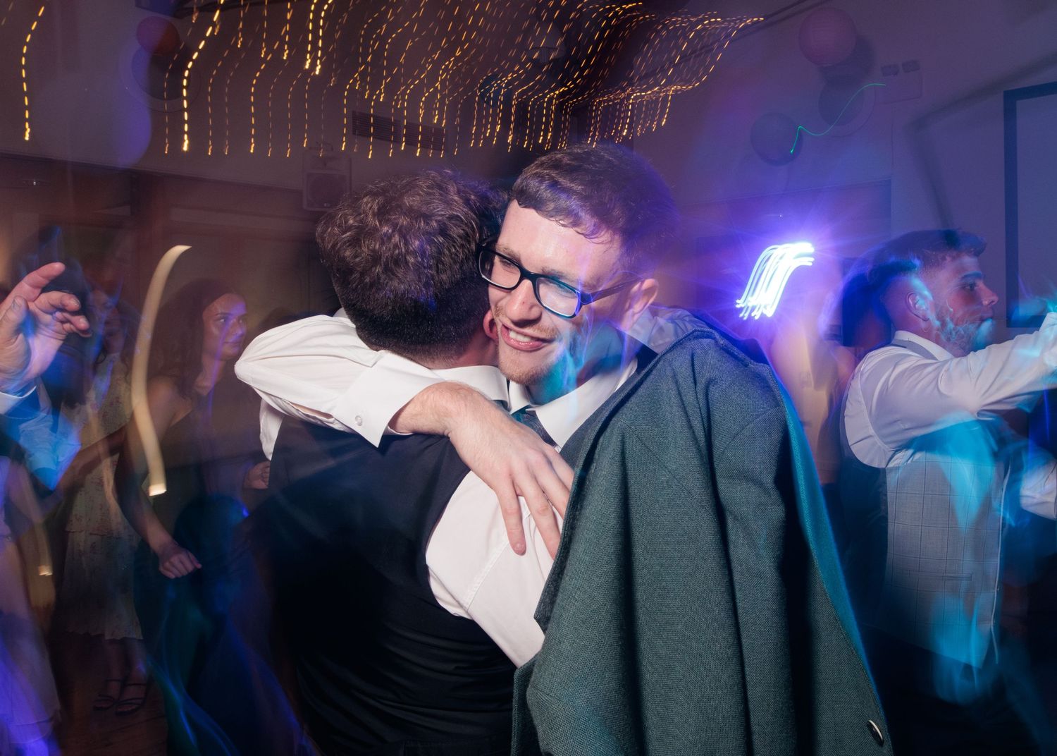 A couple shares a tender dance embrace on a dimly lit dance floor with blue lighting and other dancers in the background.