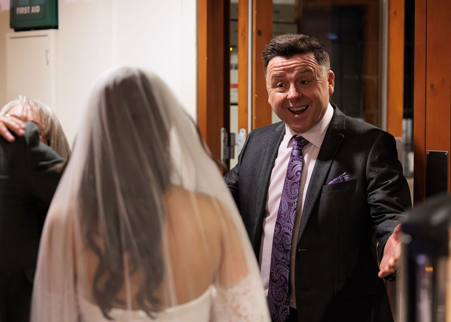 A person in a purple tie and dark suit greets a bride wearing a white veil during a wedding ceremony.