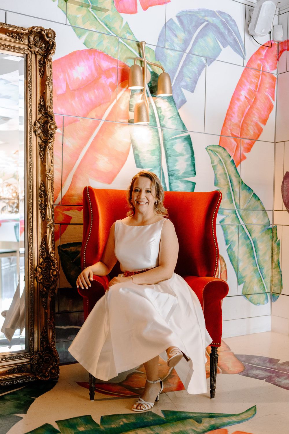 A bright interior space features tropical leaf murals, a red velvet chair, and an ornate gold-framed mirror.
