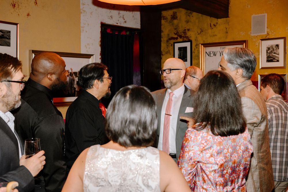 A group of people mingling at an indoor social gathering with warm lighting and framed artwork on the walls.