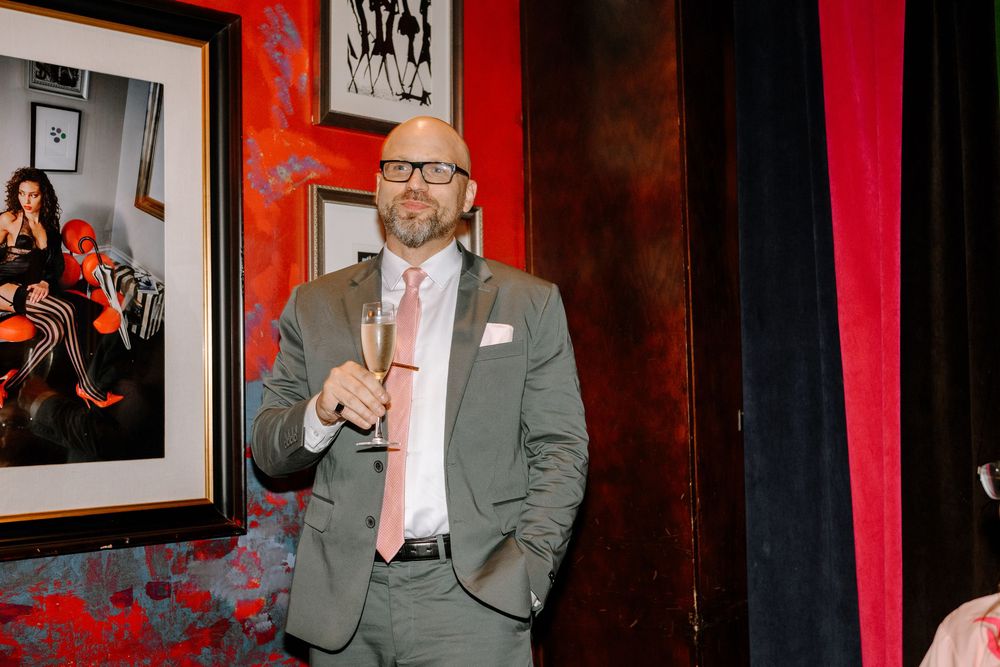 A person in a gray suit jacket and pink tie stands in front of a red curtain at an event venue.