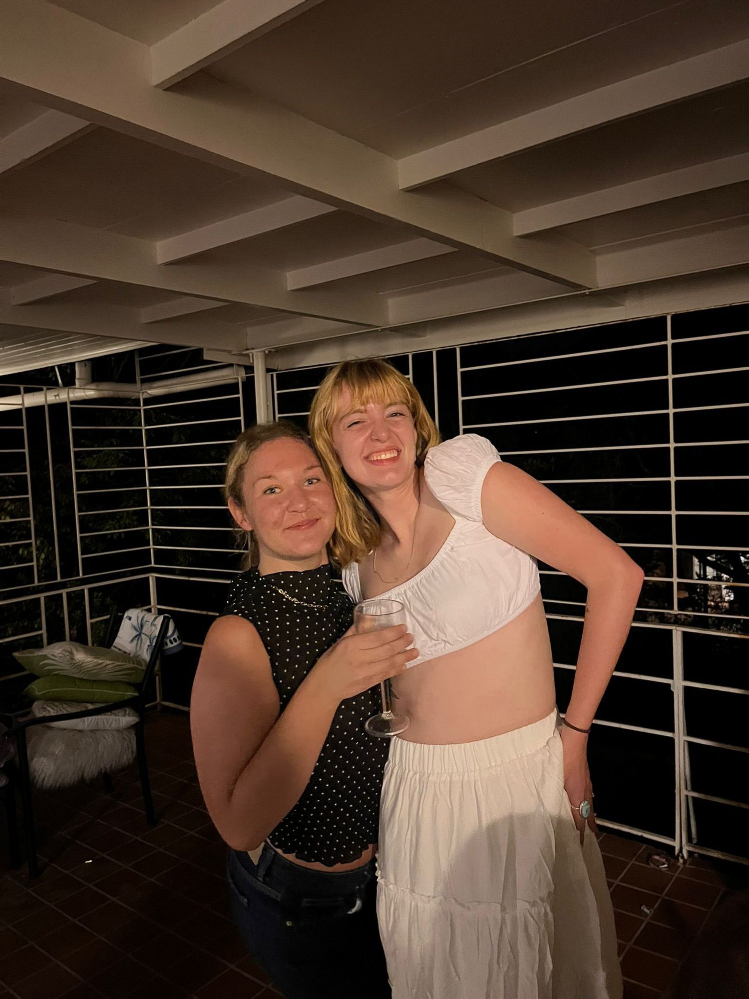 Two people laughing and hugging together in a dimly lit room with window blinds visible in the background.