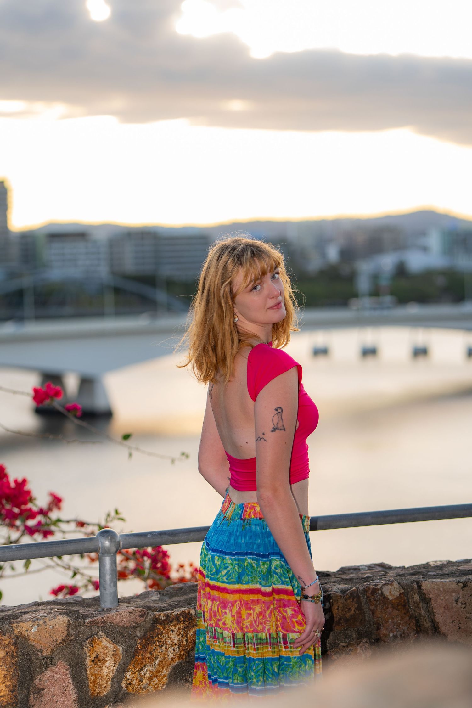 Person in pink top and colorful patterned skirt poses against sunset cityscape backdrop at urban viewpoint.
