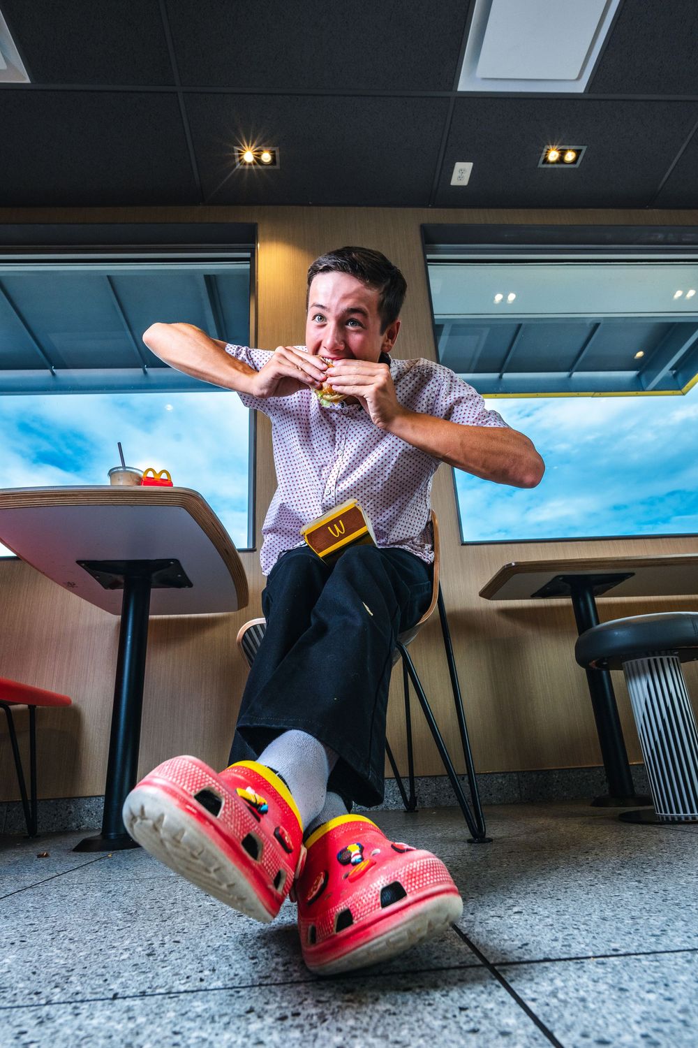 Someone eating at a restaurant table while wearing bright pink Crocs shoes, photographed from a low angle.
