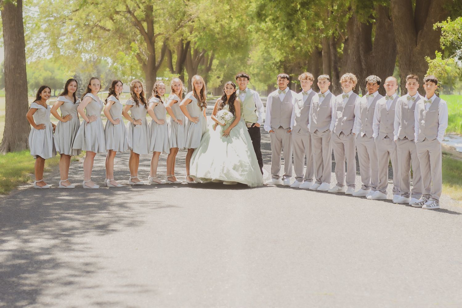 Wedding party photo showing bridesmaids in gray dresses and groomsmen in light suits on a tree-lined path.