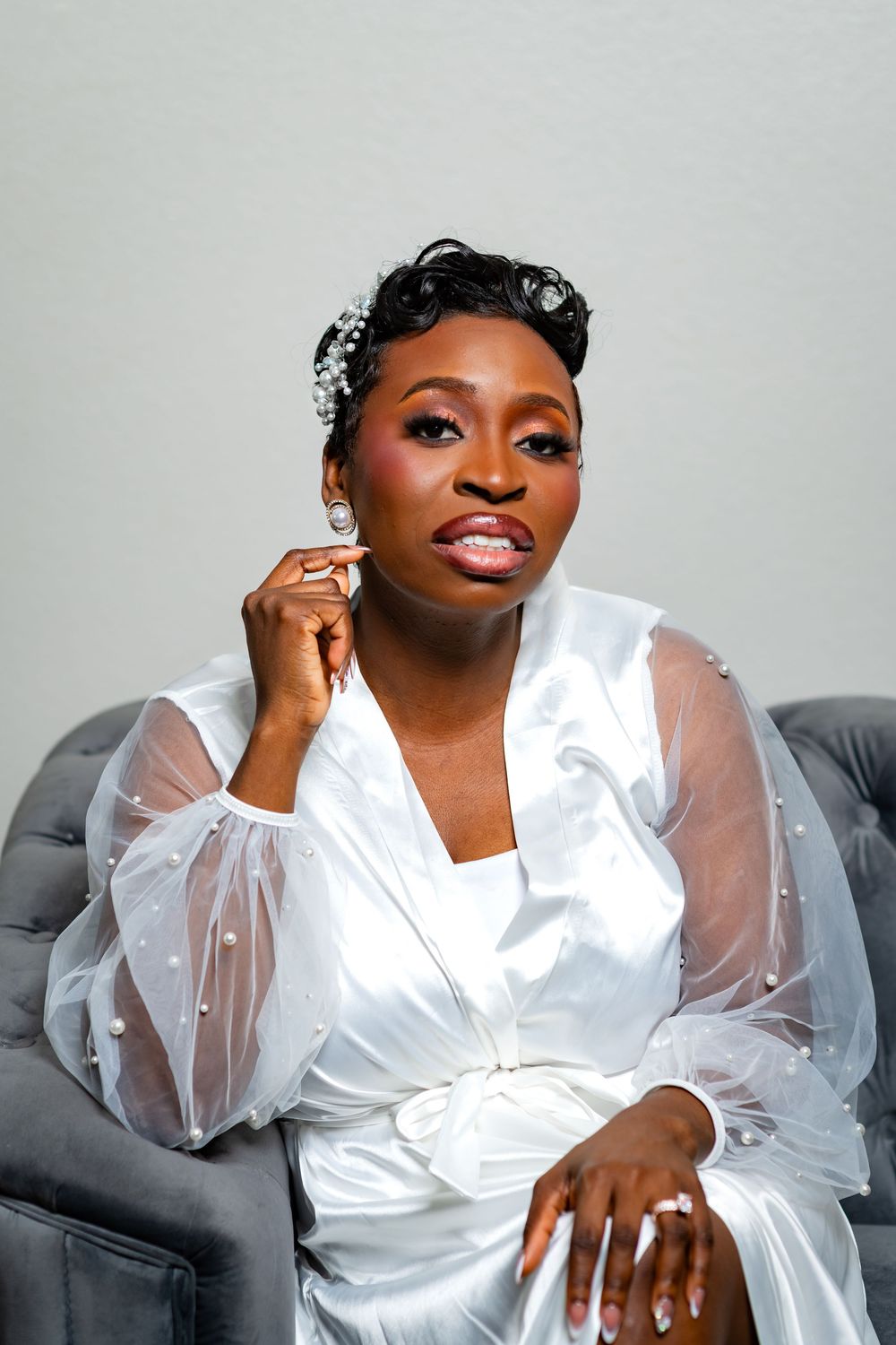 Elegant bridal preparation scene with white sheer robe and glamorous makeup on grey couch.