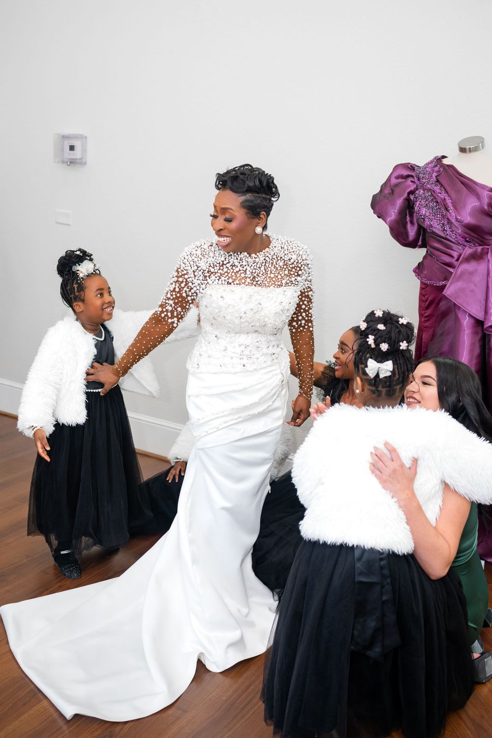 A bride in a white lace gown shares joyful moments with bridesmaids wearing black dresses during wedding preparations.