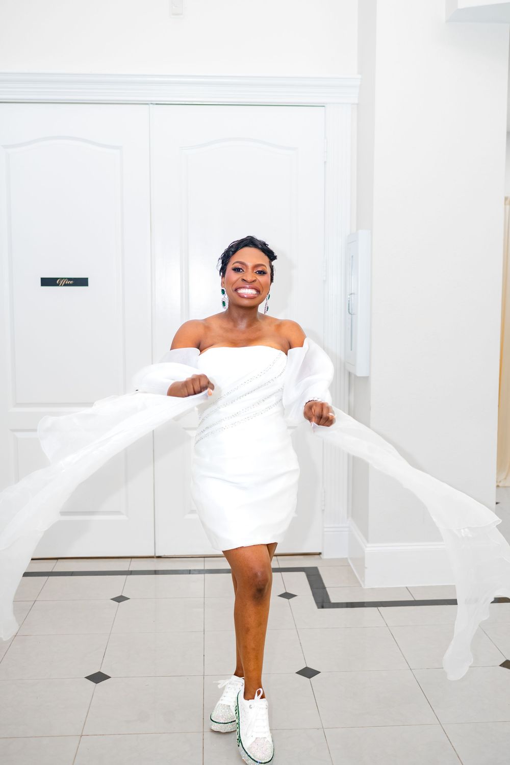 Joyful bride in short white dress and sneakers twirls fabric in bright hallway.