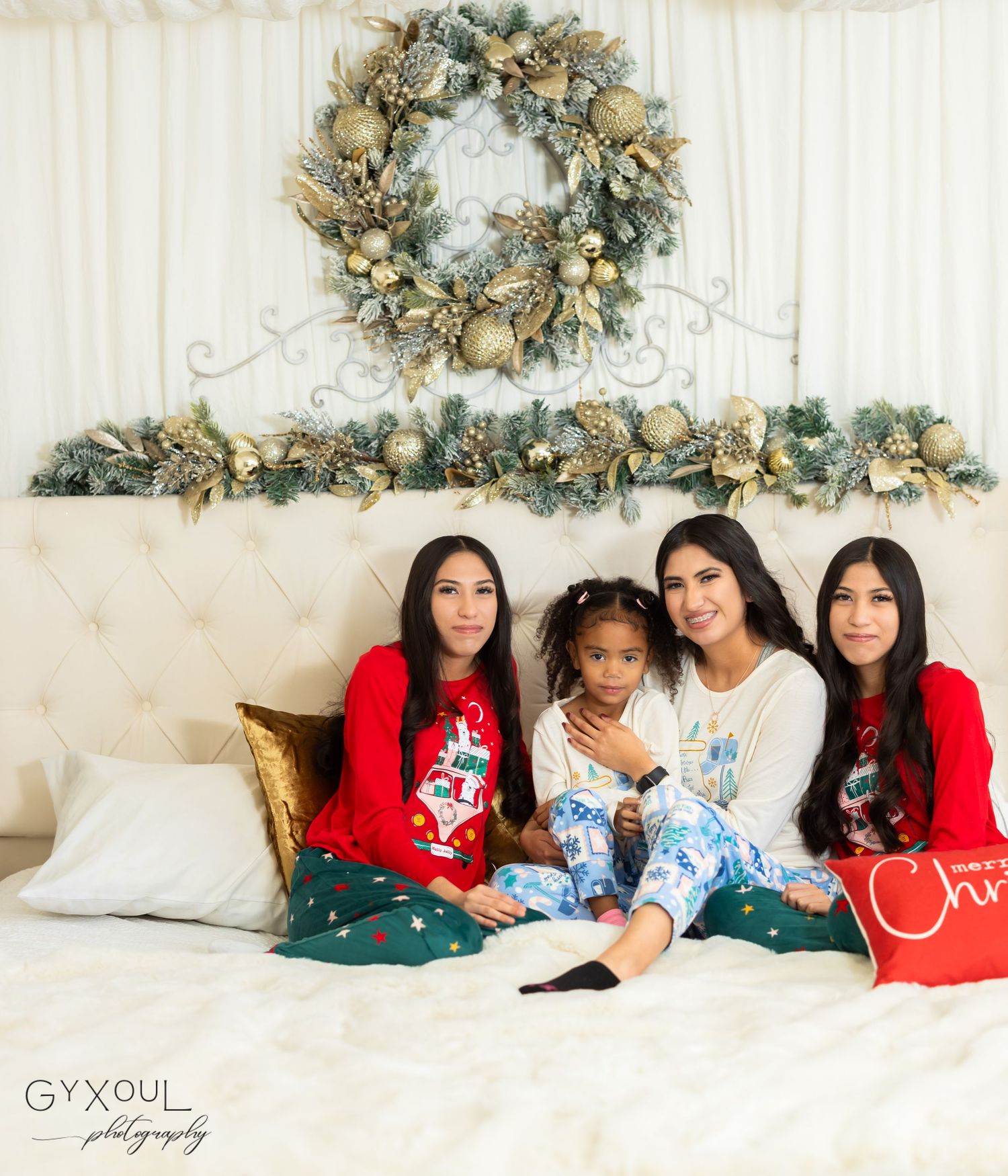 Holiday photoshoot setup with white bedding and decorative wreaths on wall in cozy bedroom setting.
