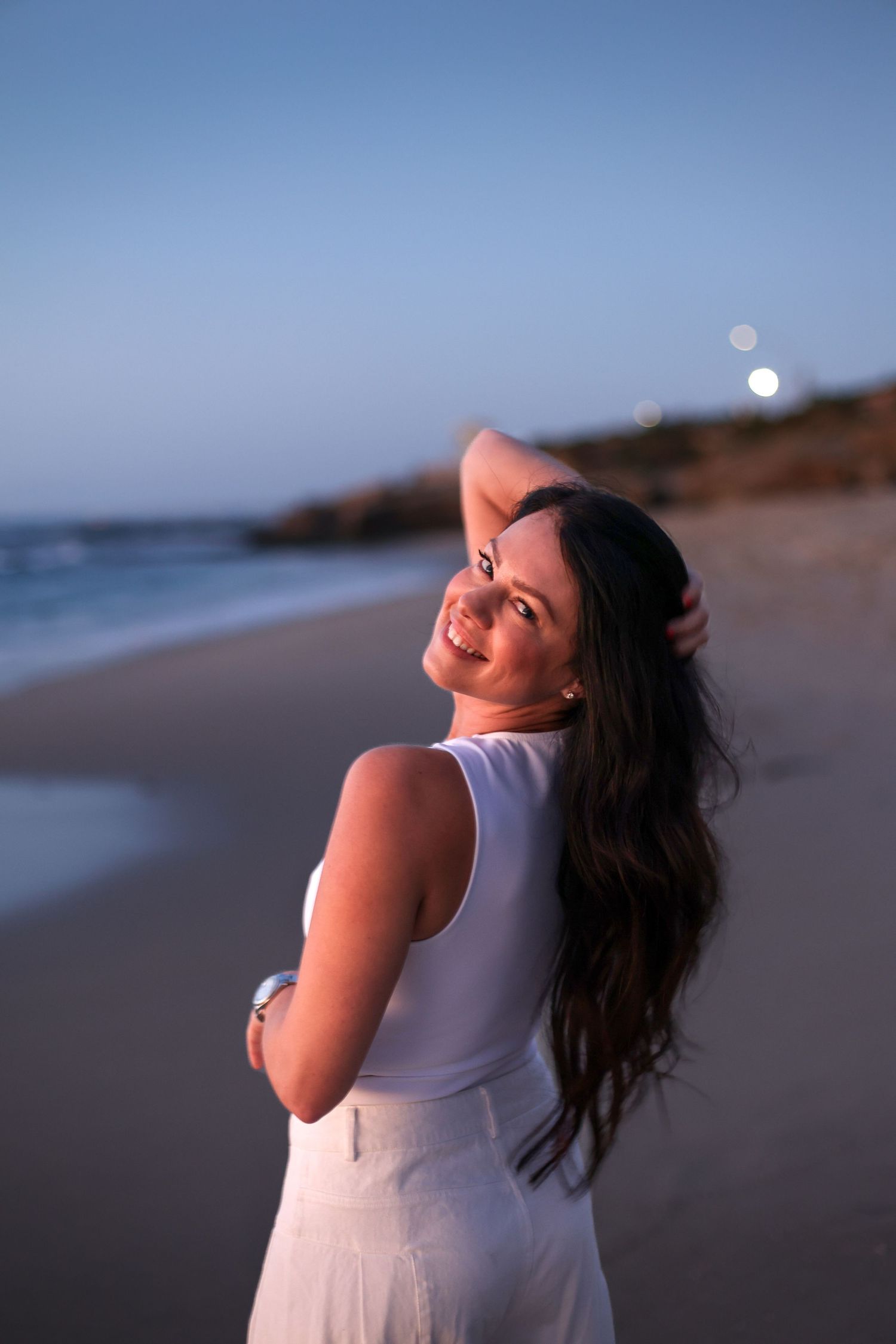 Smiling brunette woman in white dress on a sandy beach at dusk by the ocean.