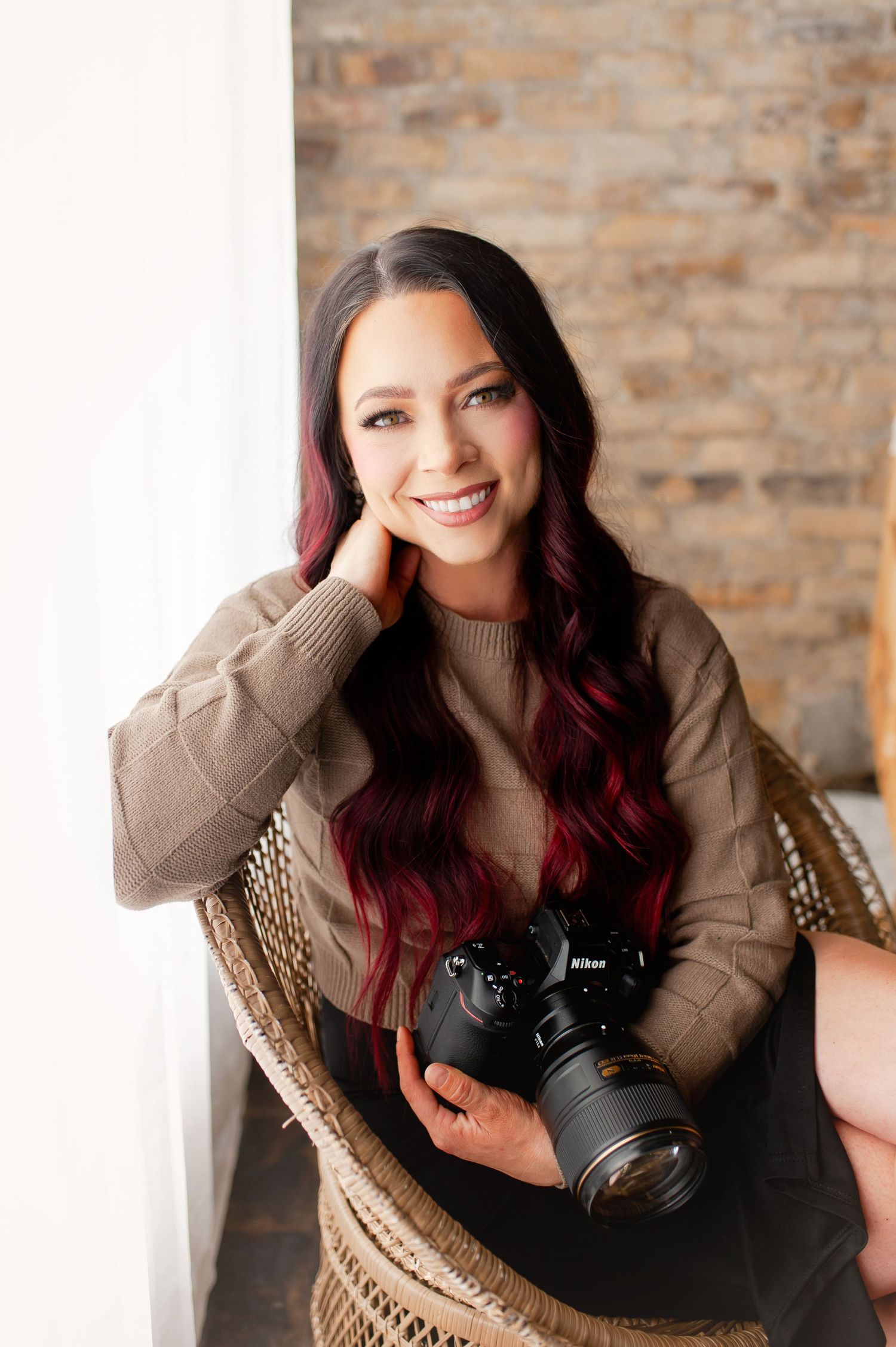 Smiling female photographer with red-highlighted dark hair holding a Nikon camera, wearing a beige sweater in a bright studio.