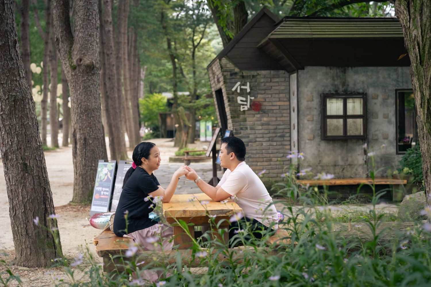 People enjoy outdoor dining at wooden tables under tall trees at a rustic restaurant patio.