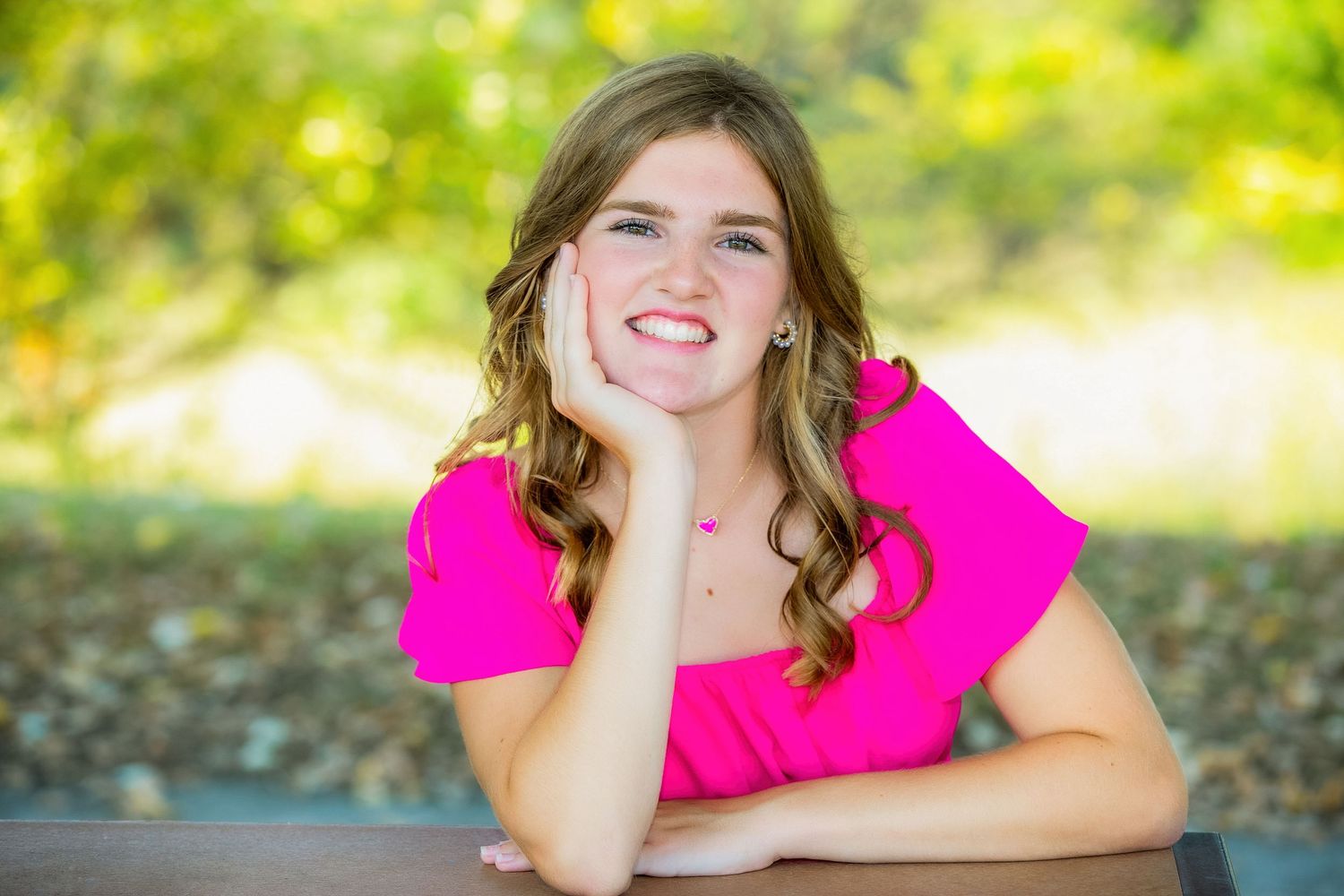 Person in bright pink dress poses playfully on autumn ground with golden sunlight and trees in background.