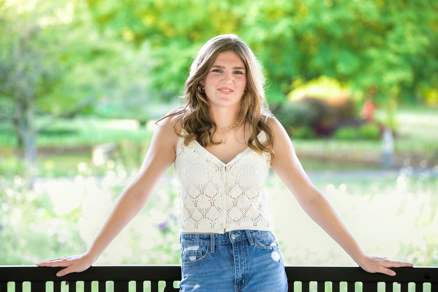 A person in casual denim sits outdoors on a sunny day surrounded by lush green foliage in a park setting.