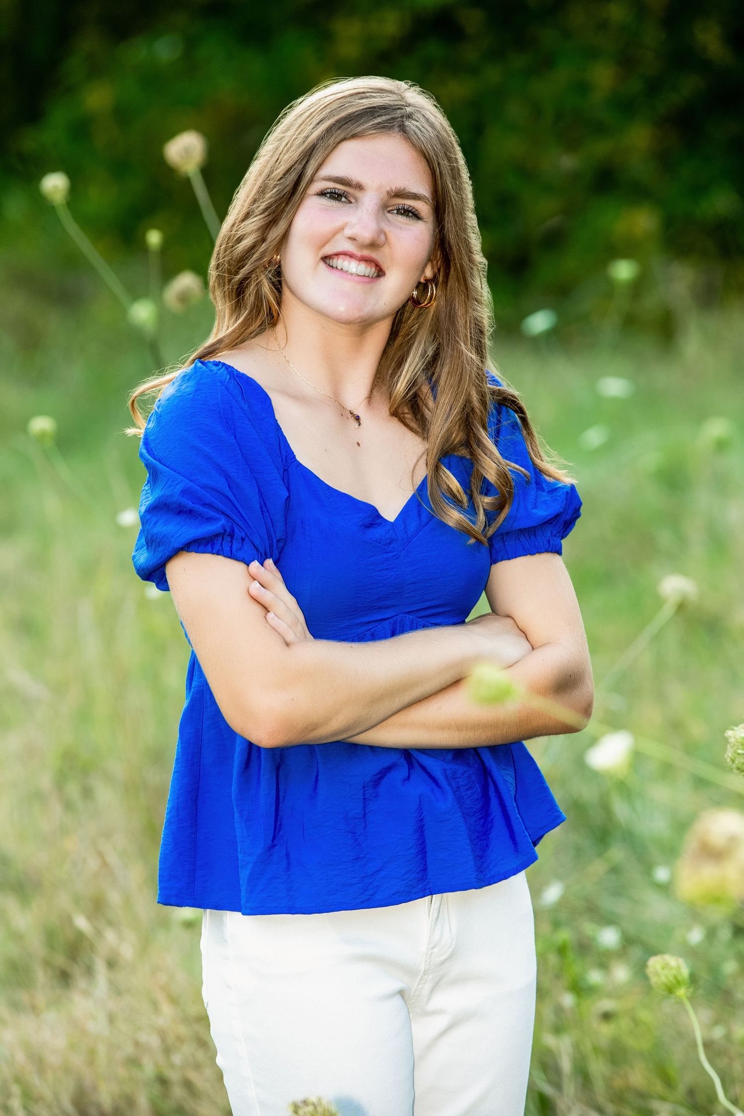 A person in a bright royal blue top and white pants poses outdoors in a grassy field with wildflowers.
