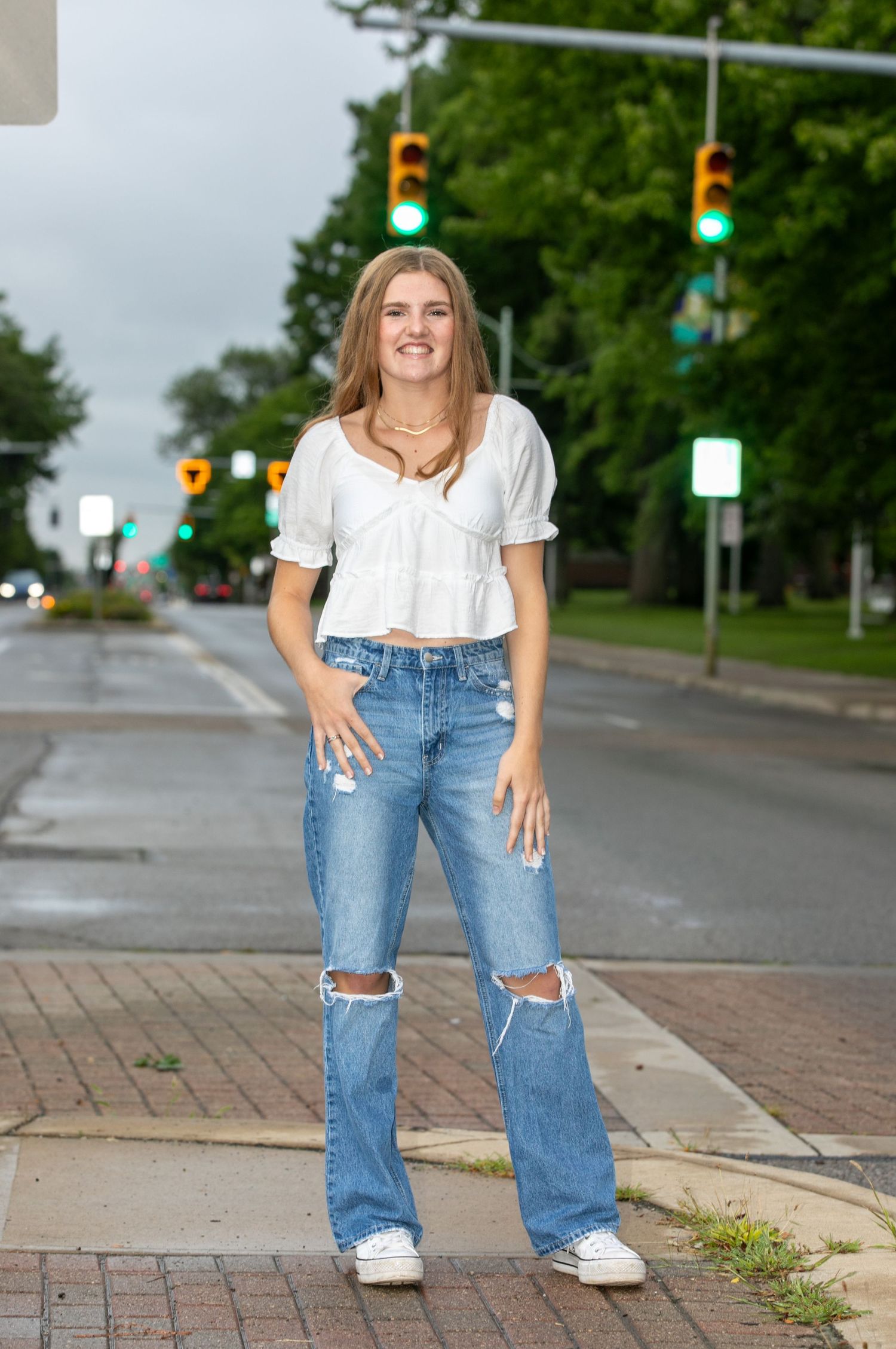 Person wearing a white top and ripped jeans poses on a brick sidewalk with traffic lights in the background.