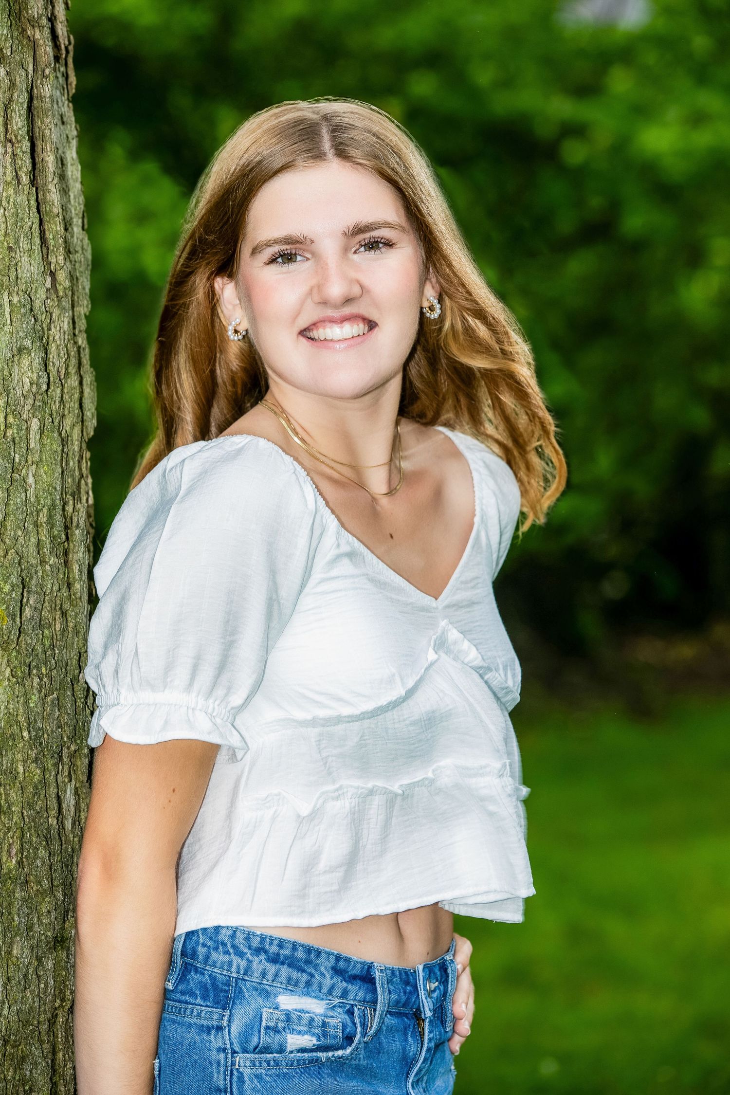 A person in a white cropped top and blue jeans poses outdoors against a green natural background.