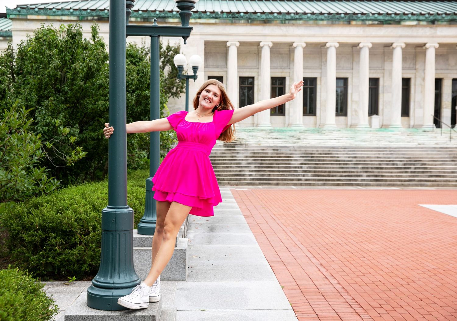 A person in a bright pink dress poses gracefully on a brick terrace with white columns and greenery in the background.
