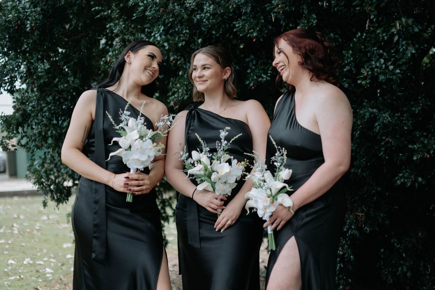 Wedding party in black dresses holding white bouquets pose together in an outdoor garden setting.