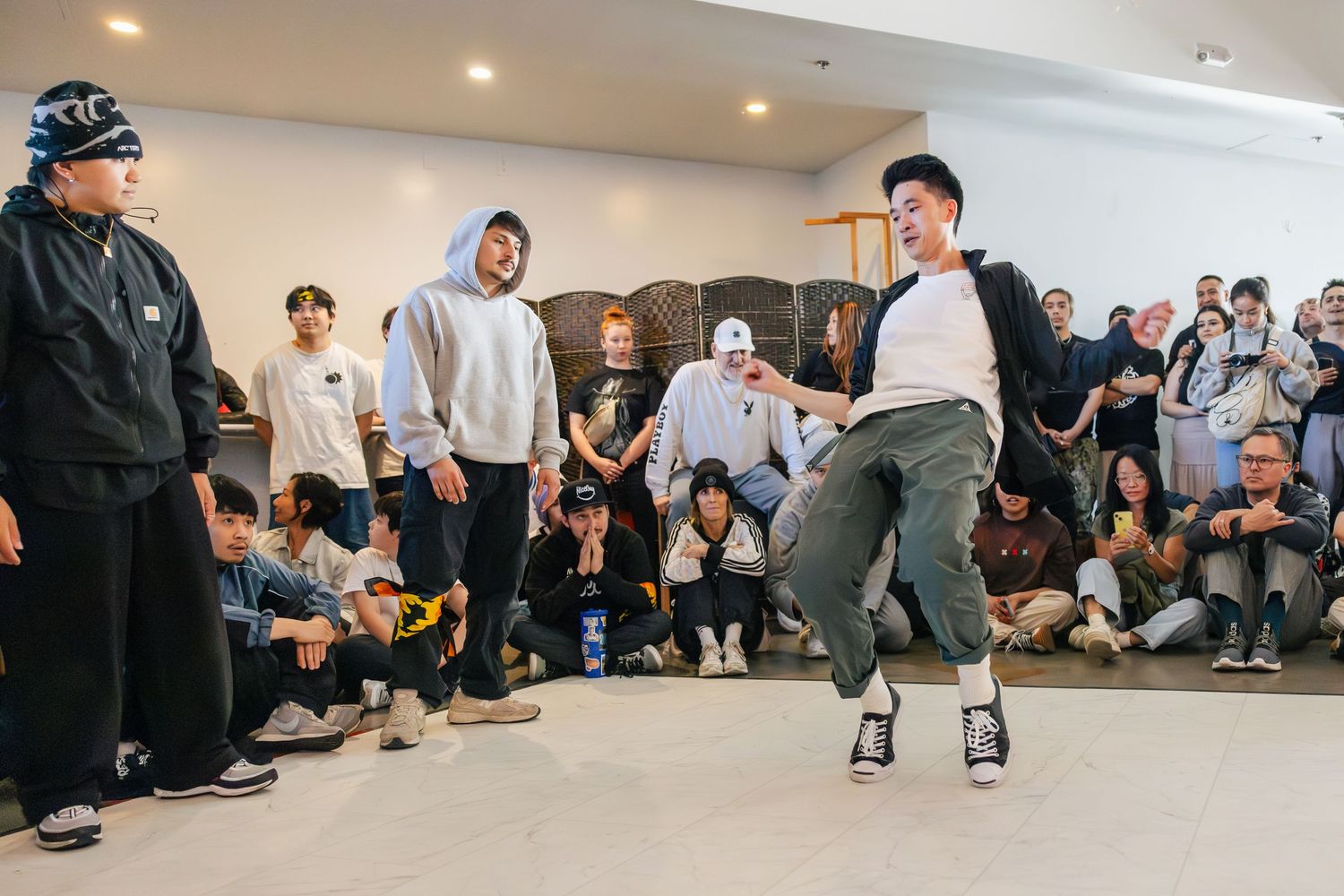 Dancers practicing breakdancing moves on a studio floor with spectators watching from the sides.
