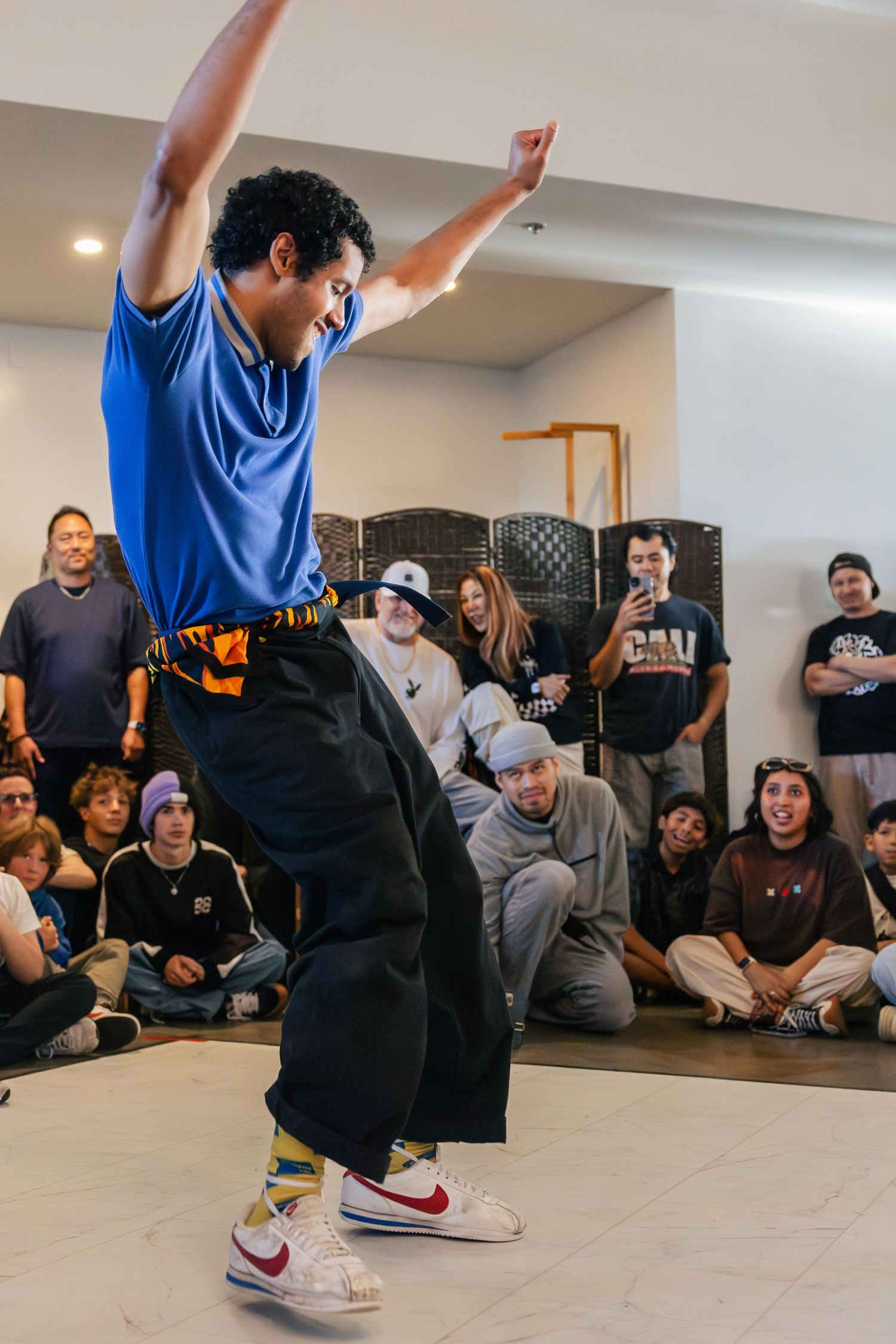 Dancer in blue shirt and black pants performs energetically in front of seated crowd at indoor dance event.