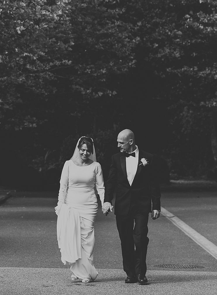 A black and white wedding photo of a couple walking together outdoors with trees in the background.