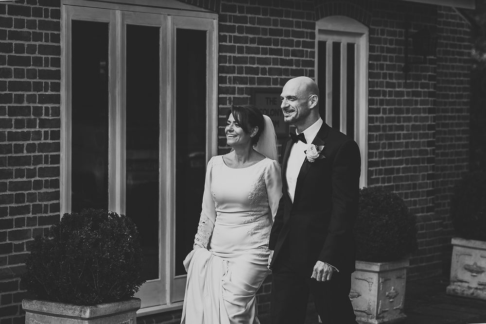 Couple poses together in formal attire against brick wall during wedding photoshoot.