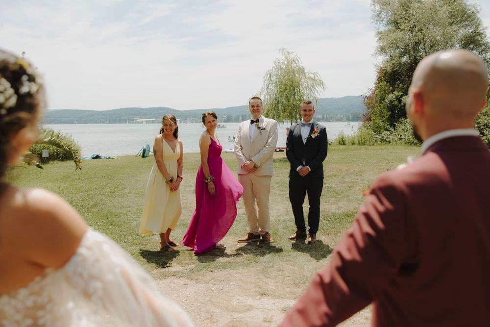 A wedding ceremony takes place on a scenic lakeside beach with guests gathered on the sand and water in the background.