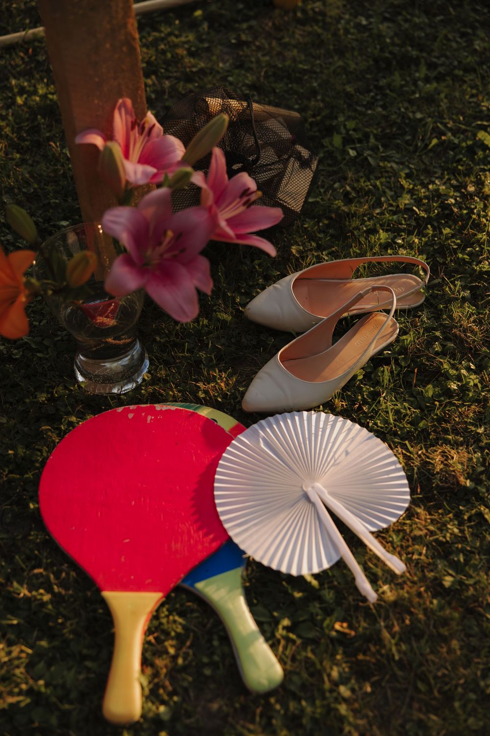 Pink lilies in a vase with ballet flats, ping pong paddles and a decorative paper fan arranged on grass in warm sunlight.