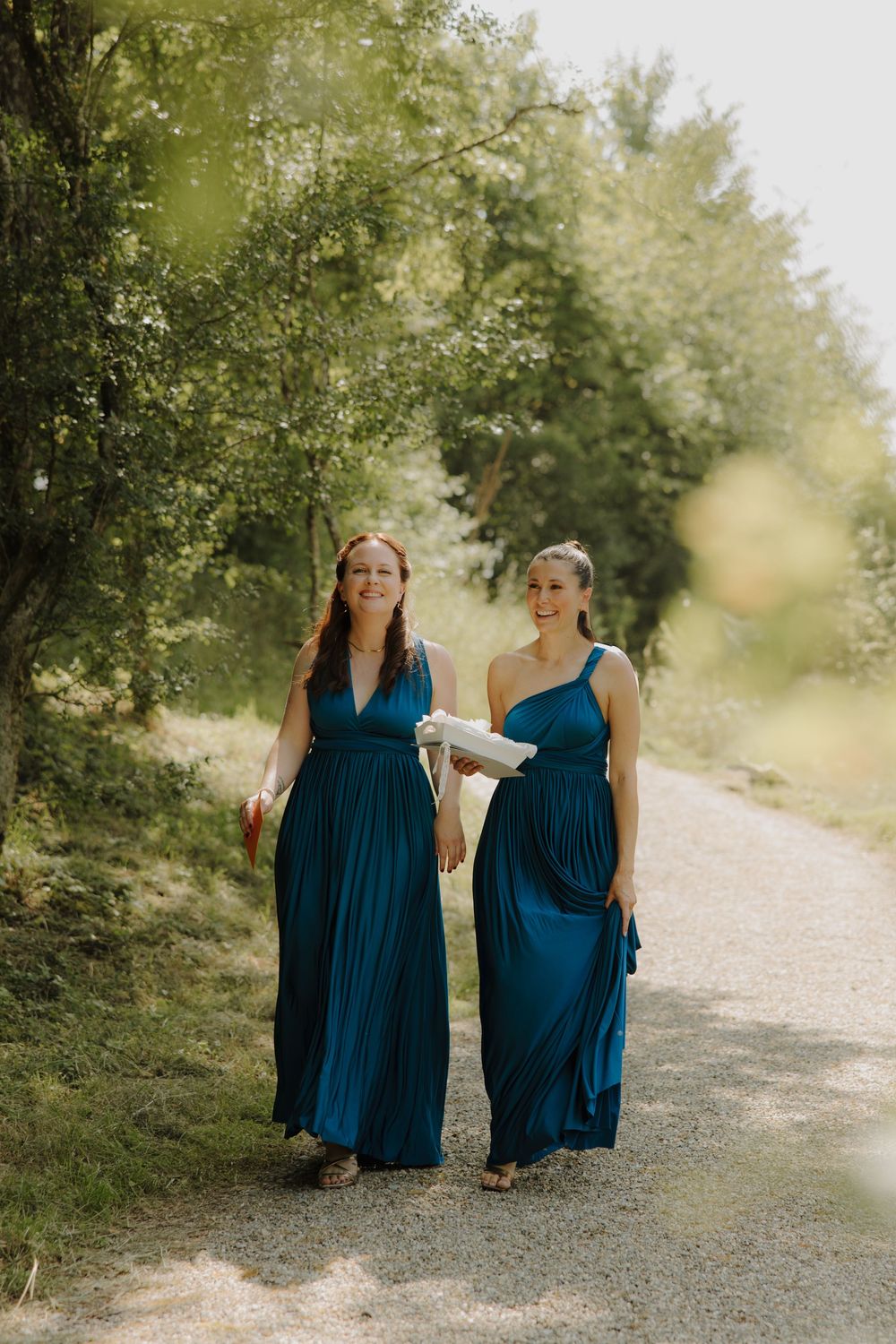 Two people in elegant navy blue dresses walk together on a gravel path surrounded by lush green trees.