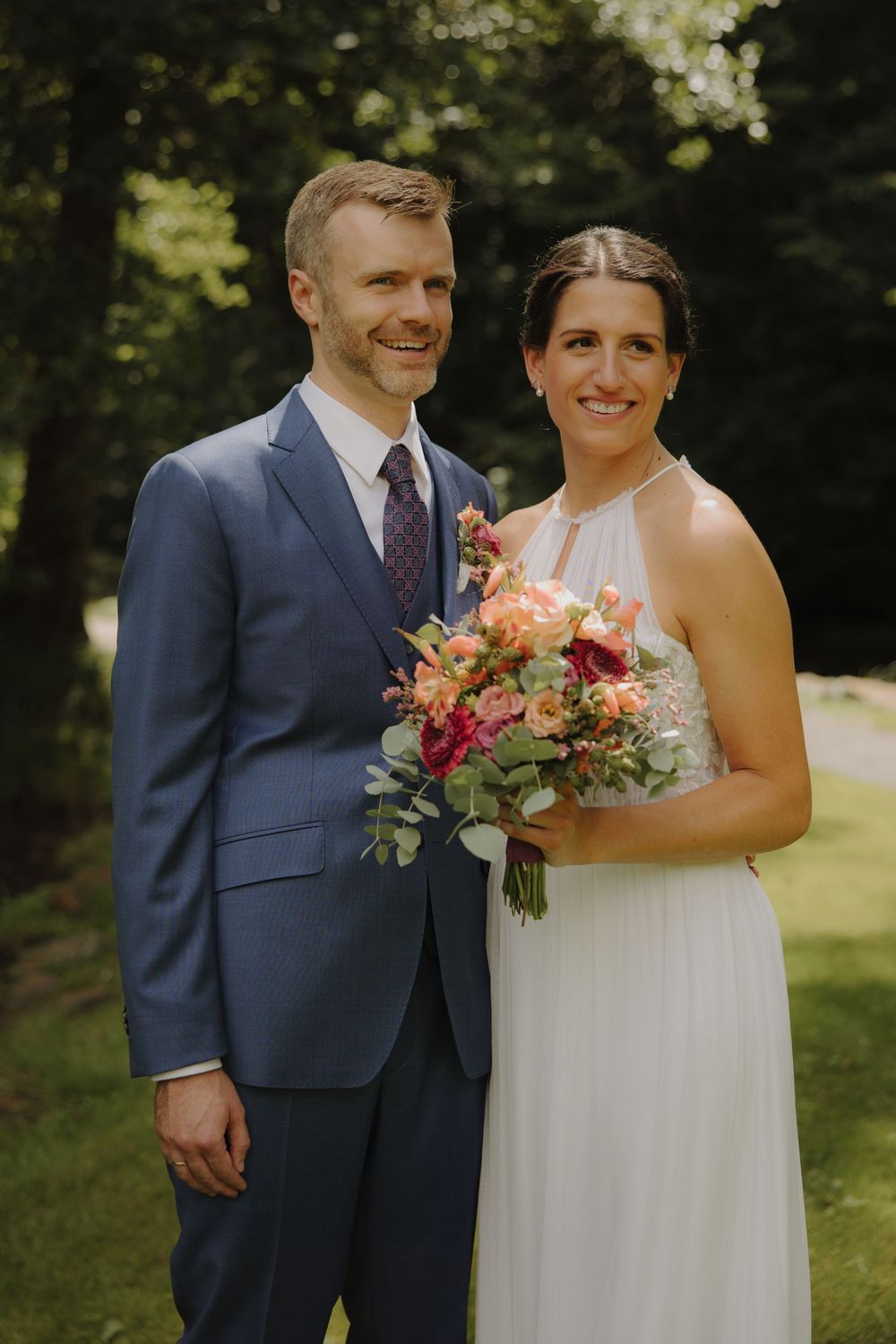 A couple in wedding attire share a joyful moment outdoors, with the bride holding a pink and coral floral bouquet.