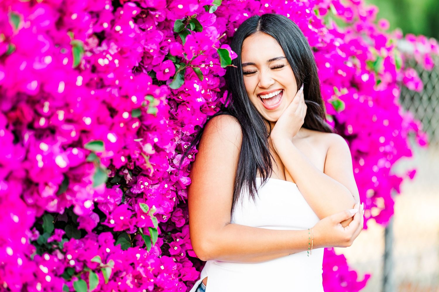 Someone in a white dress laughs joyfully against a backdrop of vibrant pink bougainvillea flowers in bloom.