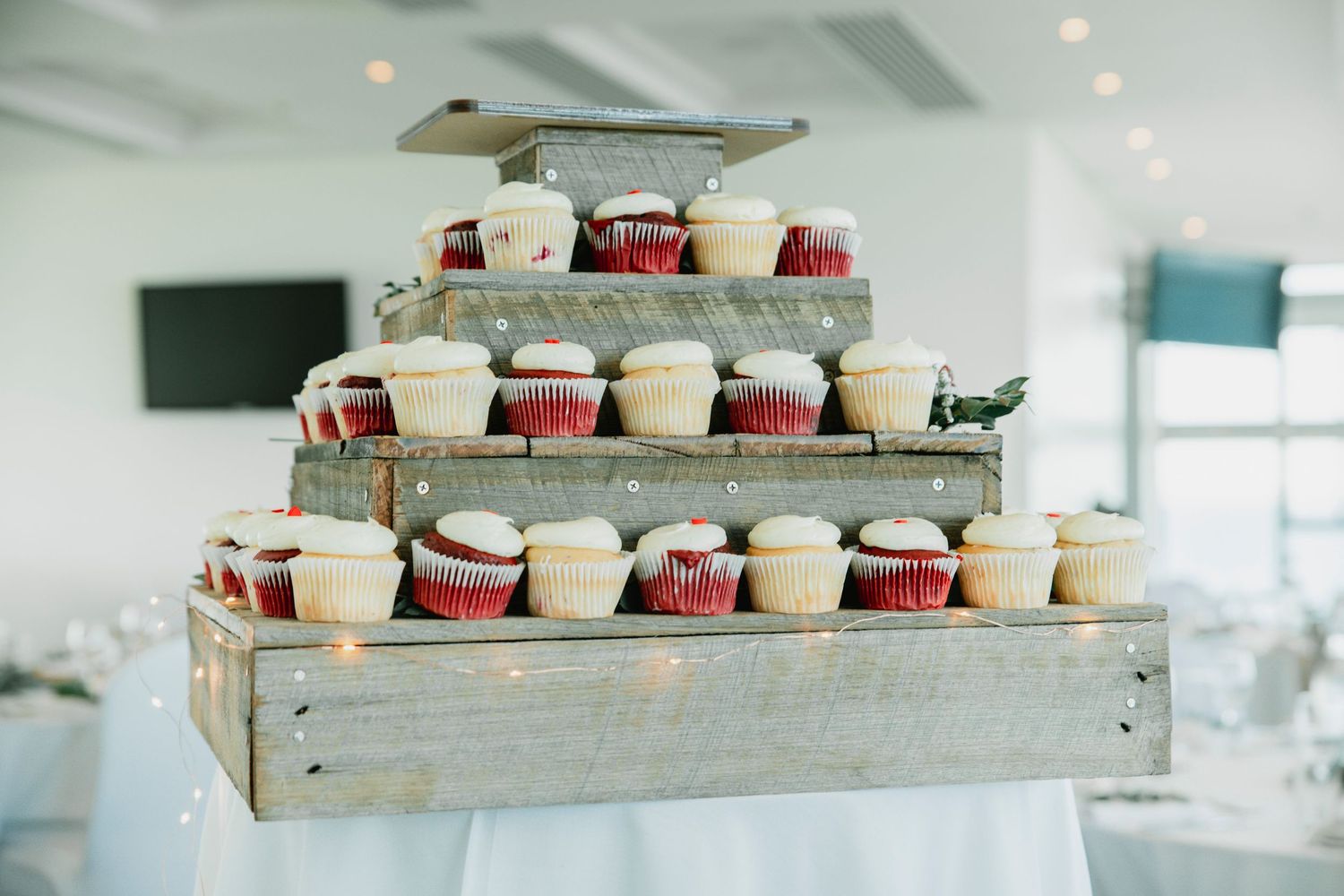 Rustic wooden tiers display white and red cupcakes arranged in rows at a wedding reception venue.