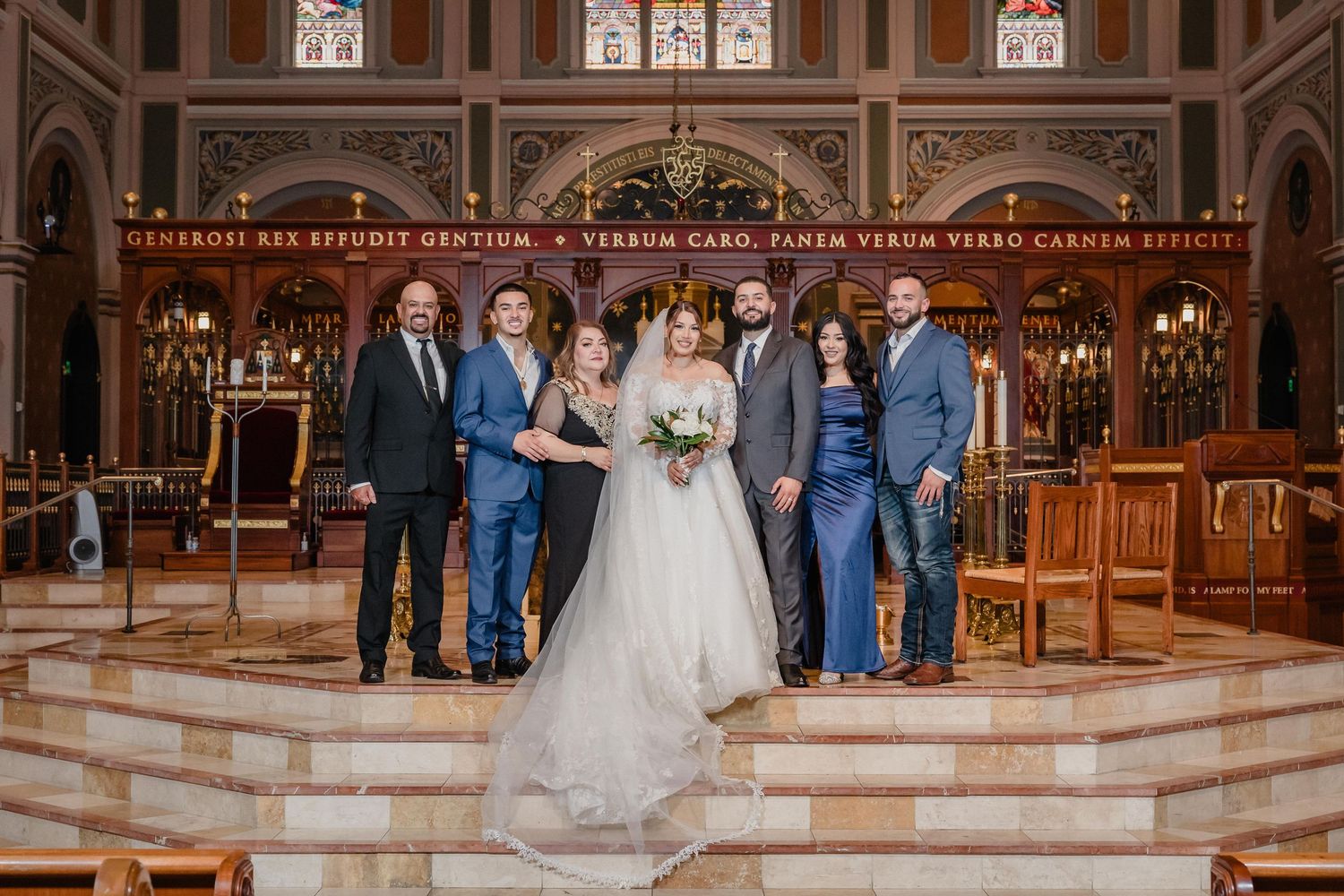 Wedding party stands in elegant church with ornate wooden pews and stained glass windows in the background.