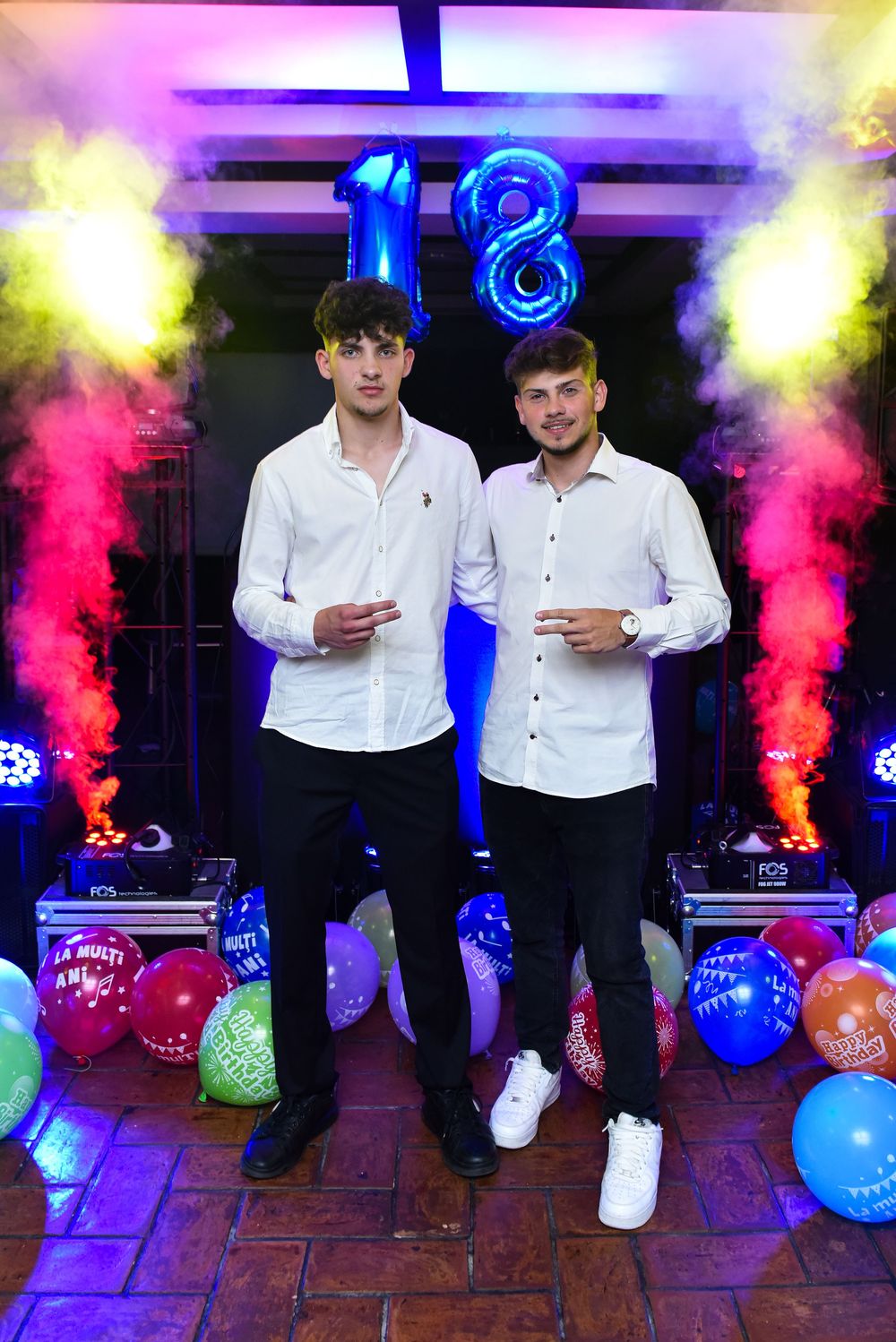 Two figures in white shirts dance against a backdrop of colorful smoke and party balloons.