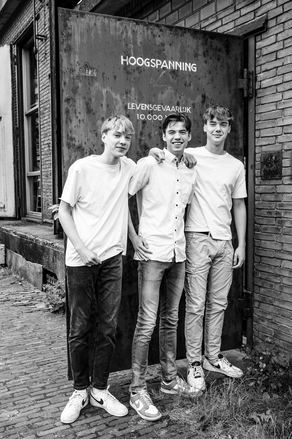 Drie broers poseren voor een oude deur met de tekst 'Hoogspanning' tijdens een fotoshoot op het Hembrugterrein