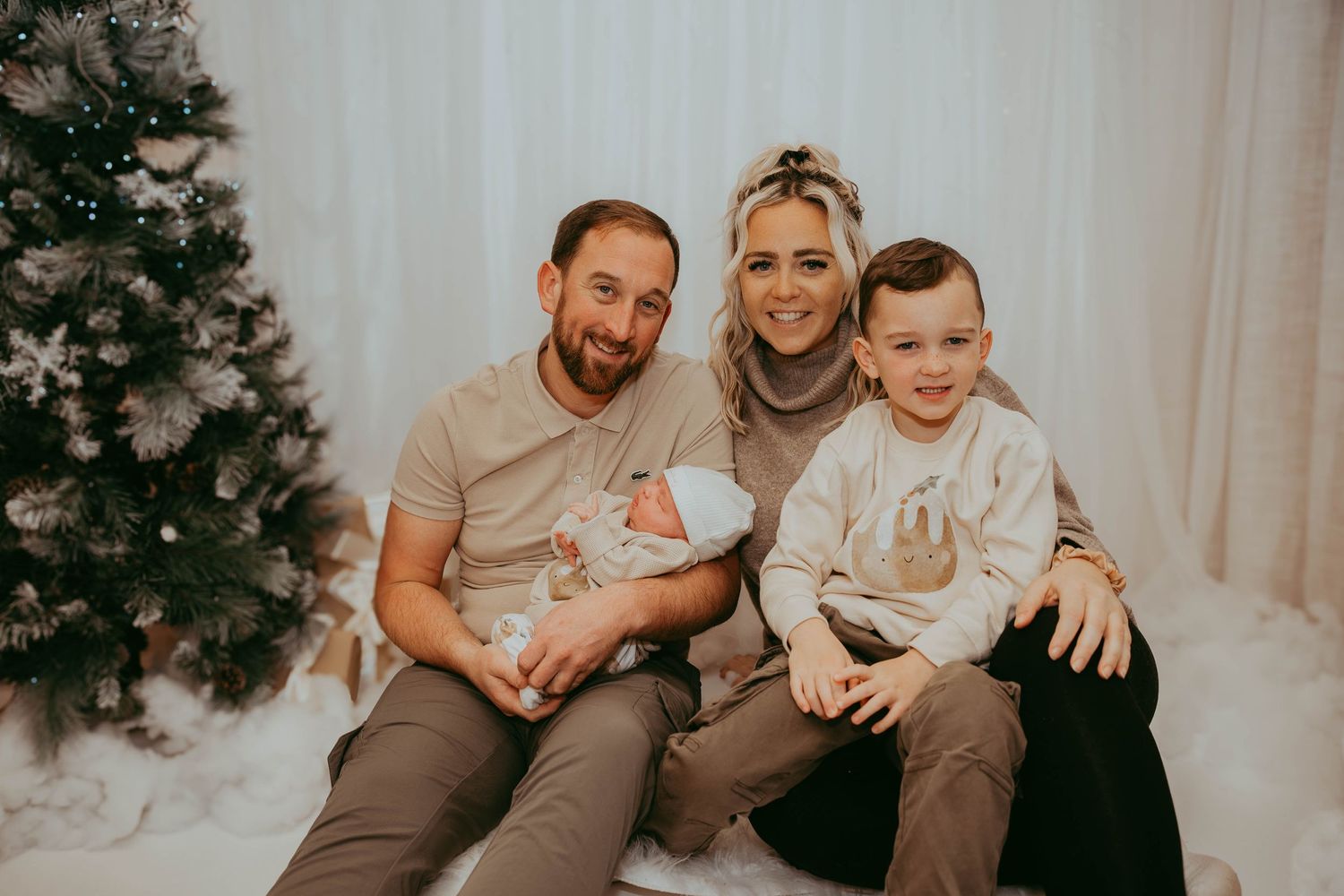 Casual family holiday portrait in neutral colors against white backdrop with Christmas tree.