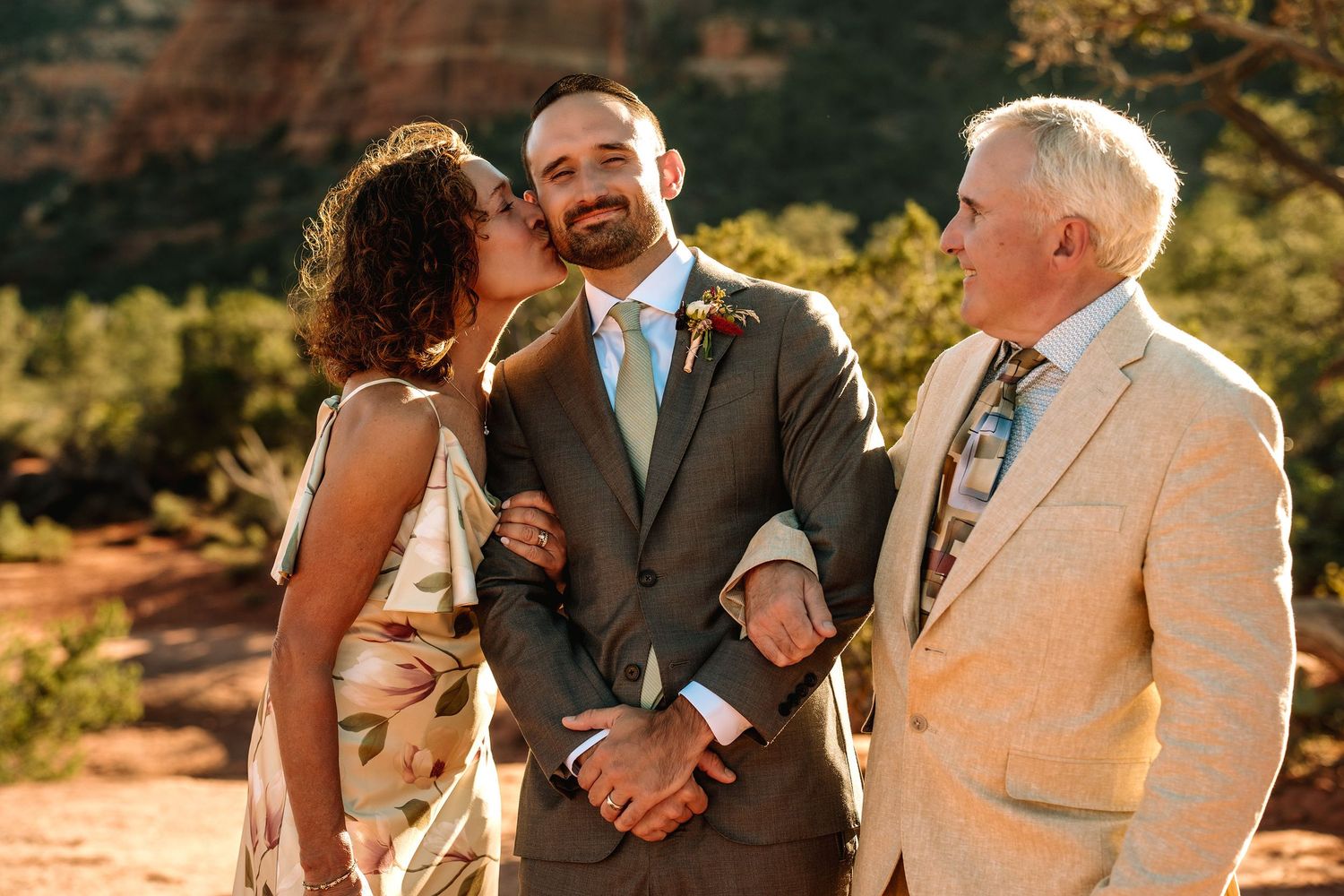 Groom in gray suit kissed on cheek by woman while older man smiles outdoors at Sedona Arizona wedding.