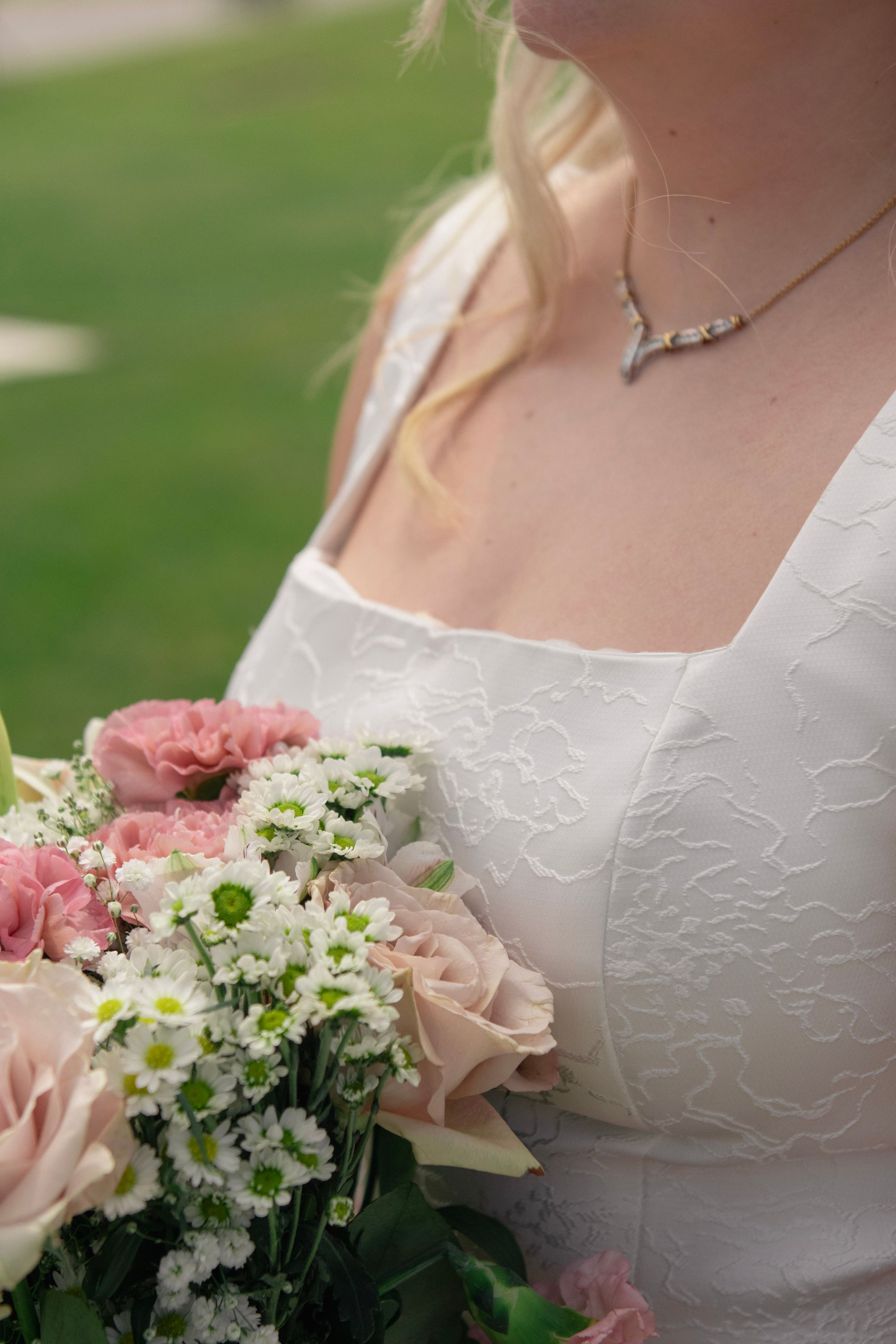 Bride in white lace dress holding pink roses and white flower bouquet, wearing silver necklace, outdoor wedding photo.