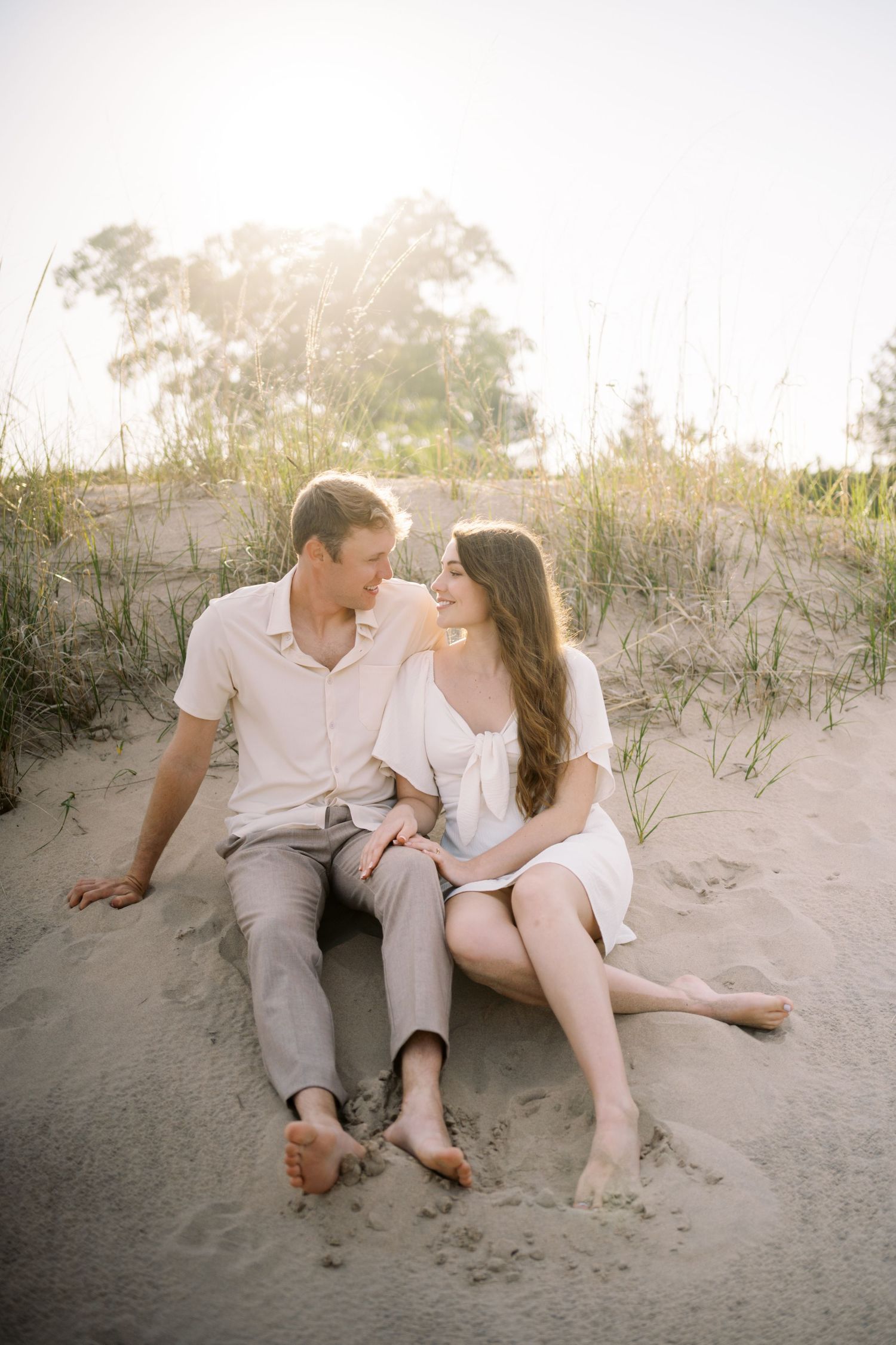 A relaxed couple in white outfits sits together on a sandy beach at sunset with dune grass in the background.